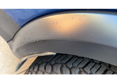 Close-up of a blue car's wheel well showing a gray bumper with a noticeable scrape along the edge.