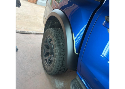 Blue pickup truck with rugged all-terrain tire and black fender flare viewed from the side, on a driveway.
