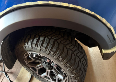 Blue vehicle's wheel well masked with tape, prepping for paint; rugged all-terrain tire and alloy wheel visible.
