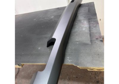 Long gray automotive trim piece with angular ends laid on a workbench in a workshop setting.