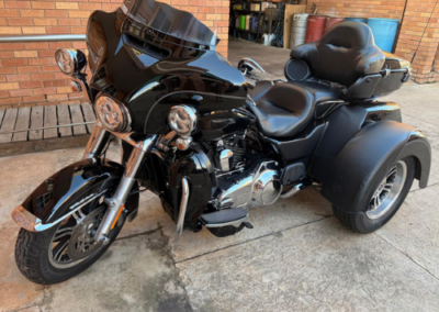 Black touring trike motorcycle with two front wheels, large windshield, and rear storage, parked on a concrete driveway beside a brick wall.
